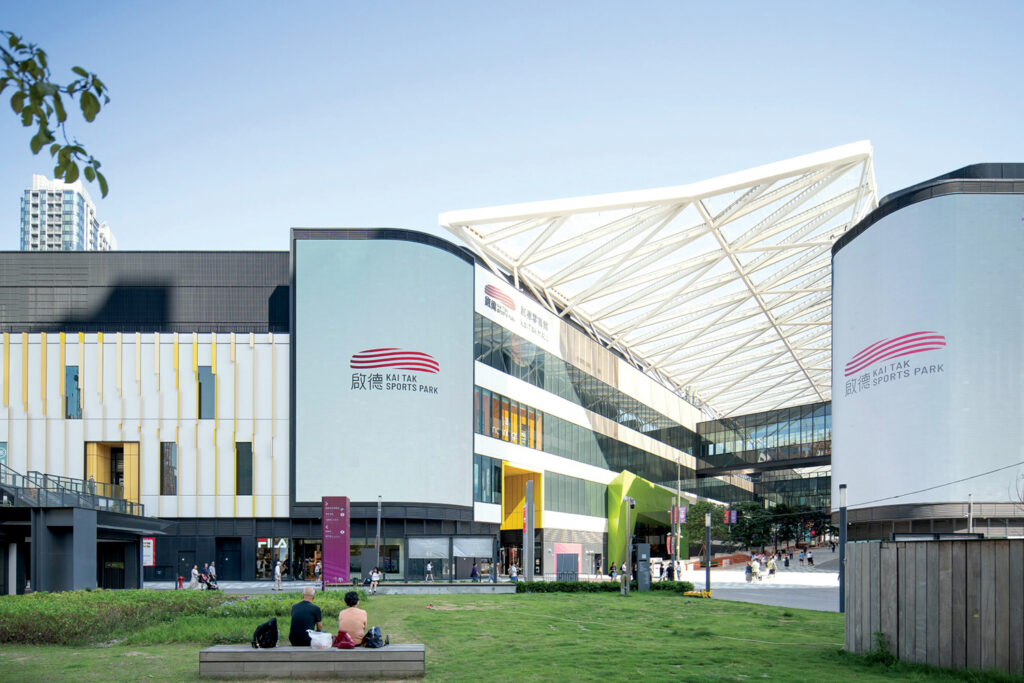 Modern sports complex with striking clear ETFE canopy, large glass windows, and vibrant colored panels. There is a green lawn in foreground with people sitting.