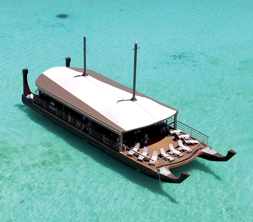 Traditional vaka boat with a curved roof, resting on clear turquoise waters, featuring lounging areas and two tall masts.
