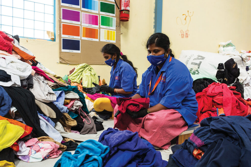Two women in blue shirts and masks sort through a large pile of colorful clothing in a brightly lit room, with a color chart on the wall.