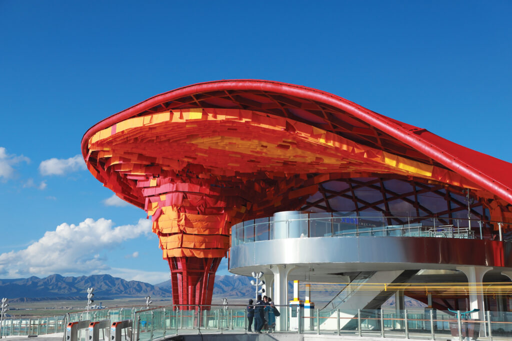 A vibrant architectural structure with a flowing red and orange roof, set against a blue sky and mountain backdrop.