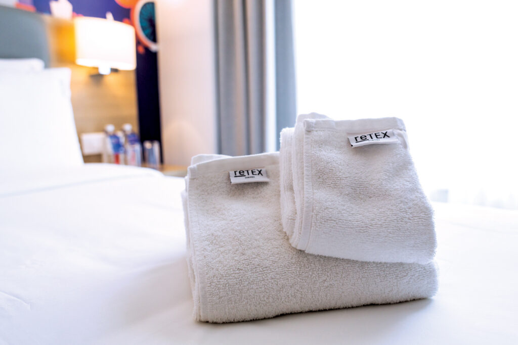 Neatly stacked white towels on a bed, featuring a light-colored blanket and a decorated wall with colorful artwork in the background.