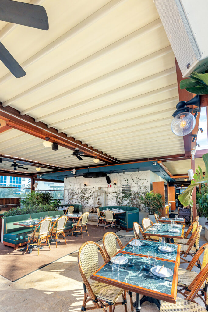 Bright, open dining area with green plants, rattan chairs and marble-top tables under a canopy; features modern decor and lighting.