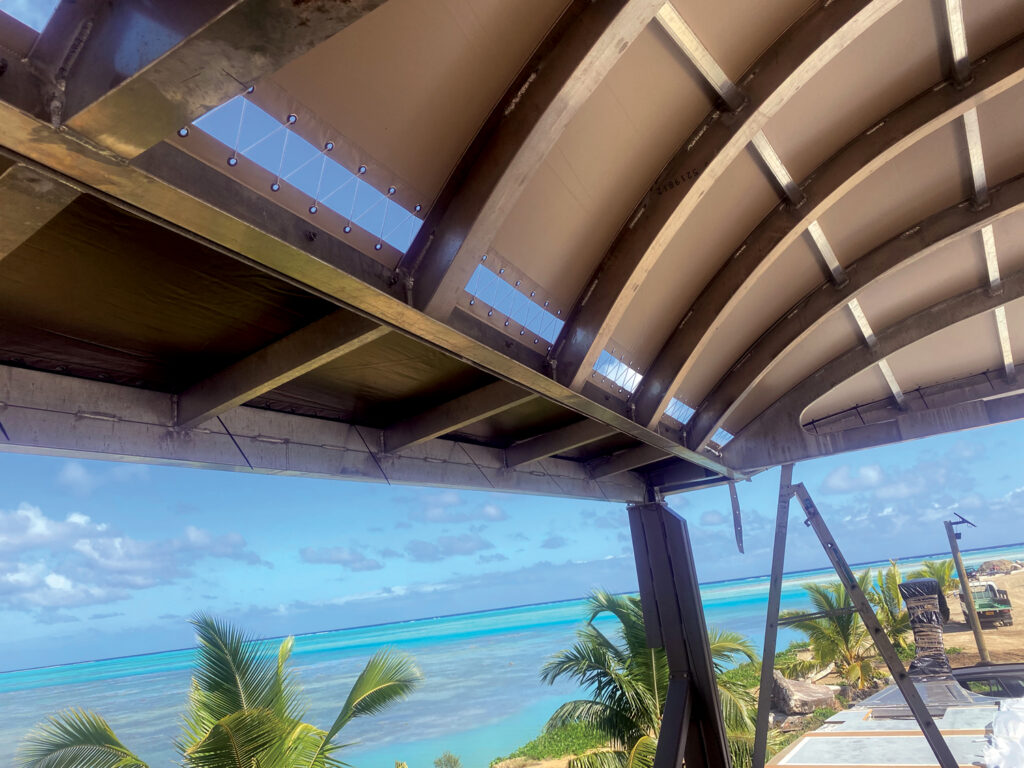 A curved metal structure frames a bright blue sky and turquoise ocean, with palm trees visible in the foreground.