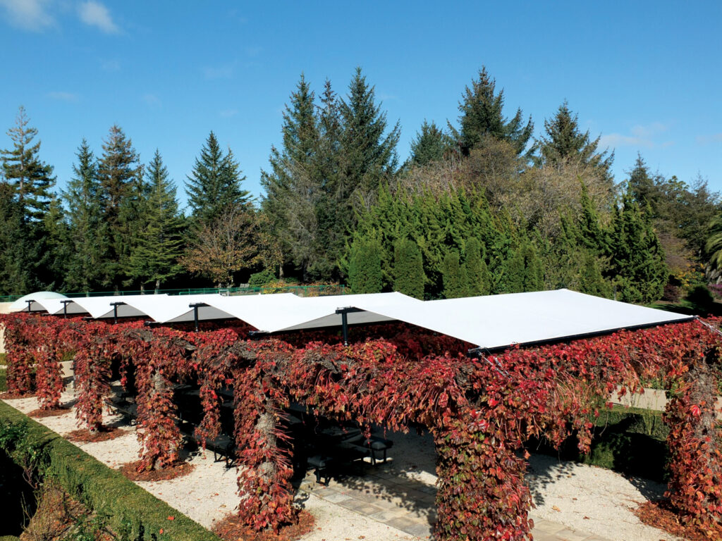 A covered outdoor space with white canopies supported by black posts, surrounded by red foliage and tall green trees under a clear blue sky.