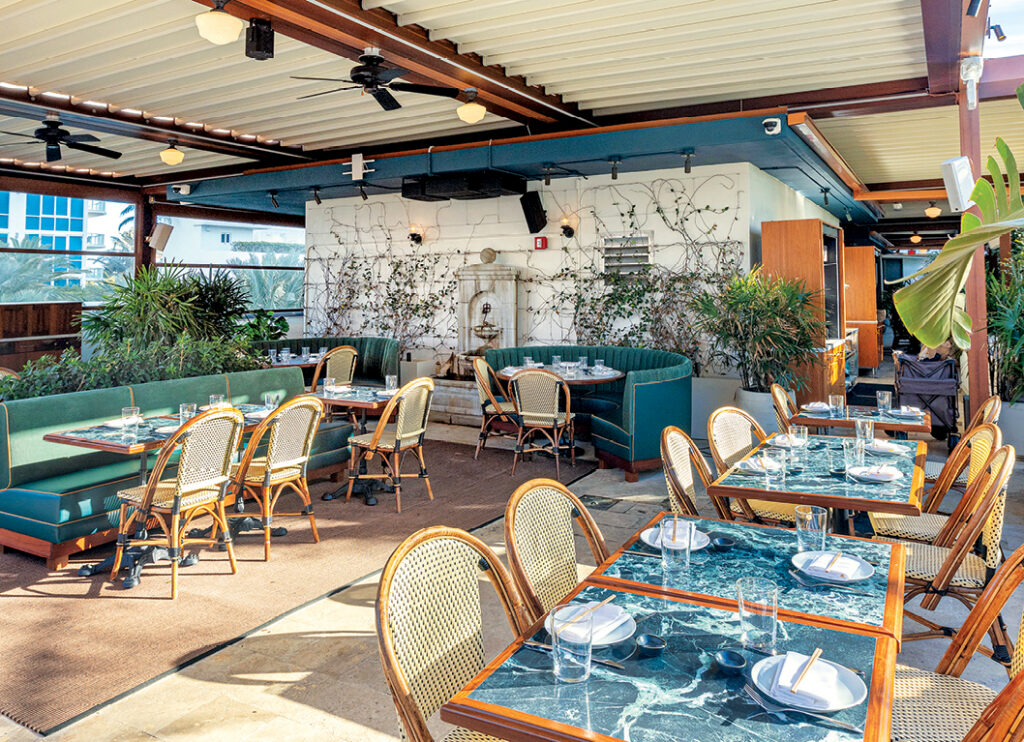 Modern outdoor restaurant scene featuring green booths, wooden tables with marble tops, and tropical plants in a bright, airy setting.