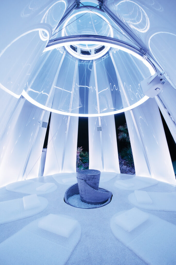 A modern, translucent sauna called Taiyo Tsubomi made with tall curved translucent ETFE walls illuminated by blue and white lights. It has a central circular reclining area and a water feature in the center.