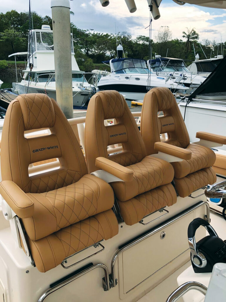 Three tan, quilted seats labeled "Grady-White" on a boat, with multiple boats visible in the background and greenery nearby.