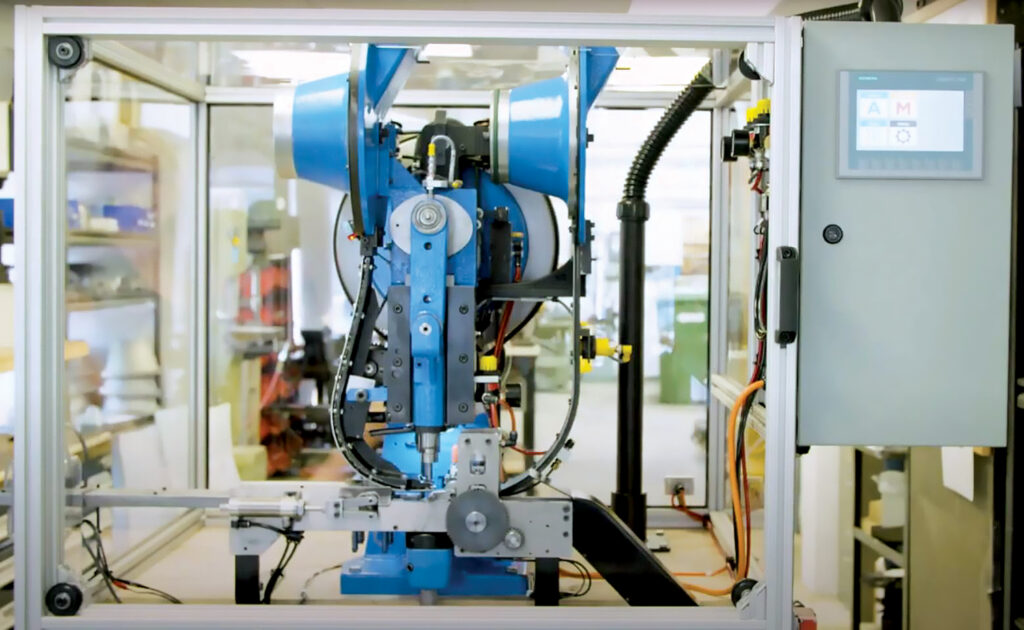 The Eagle I automatic grommeting machine from Adfast Grommeting includes a blue robotic arm and other various mechanical components housed in a transparent enclosure and surrounded by equipment and wiring.