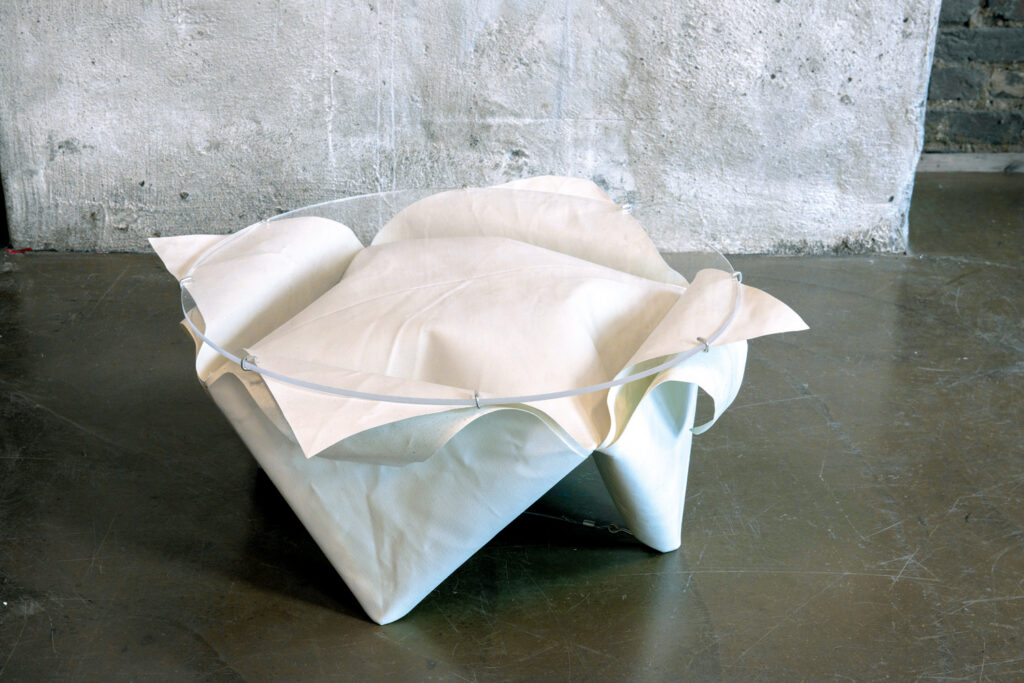 A minimalist white coffee table with soft, folded edges sits on a polished floor, complemented by a textured gray wall in the background.