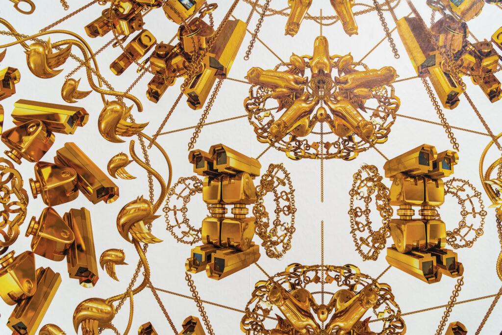 A symmetrical arrangement of intricate golden shapes that on close inspection are surveillance cameras, and chains, featuring stylized locks and floral elements against a white background. The artwork is Ai Weiwei’s graphic wallpaper The Animal That Looks Like a Llama but Is Actually an Alpaca.