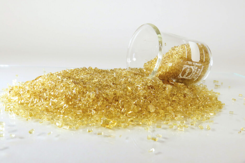 A glass beaker tipped over, spilling translucent yellow granules onto a white surface, creating a textured, shimmering effect.