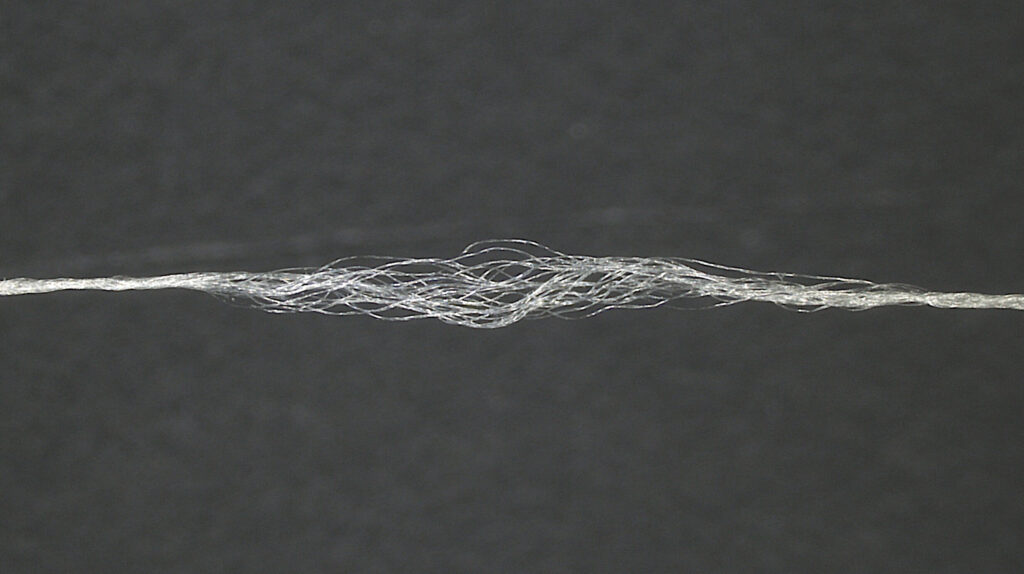 A fine, translucent yarn made of bio-based FR PEF held taught at both ends but unwound in the middle to show the fibers, set against a dark background.