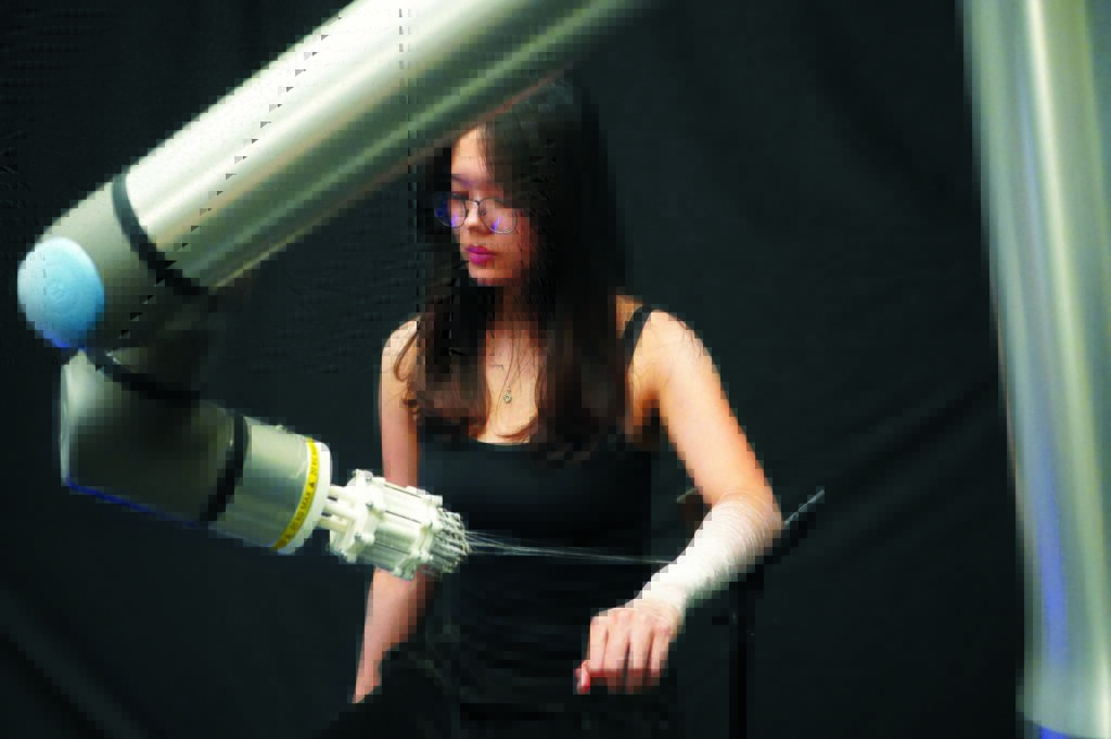 A woman with long hair sitting in a black outfit collaborates with a robotic arm, which is weaving a textile around her arm.