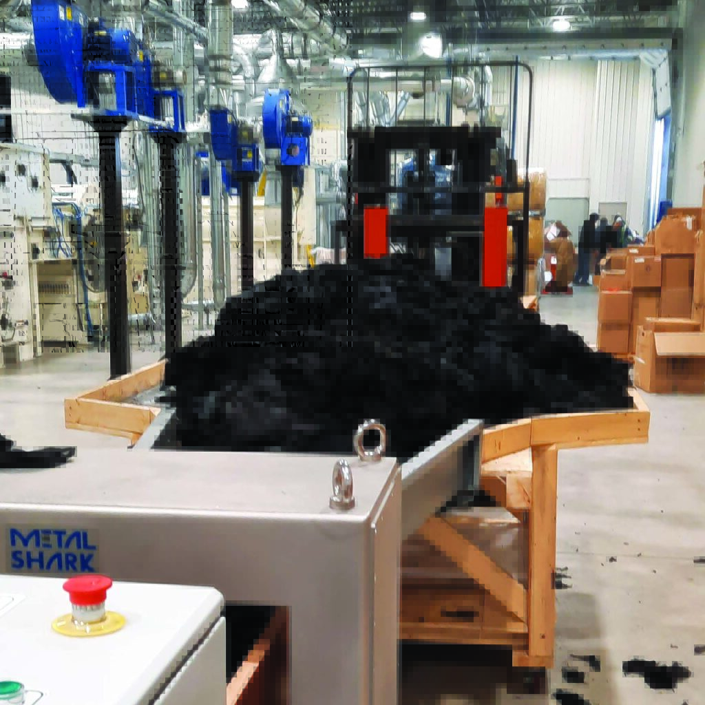 Black fabric scraps, 50% aramid, 50% FR viscose, are piled high on a platform leading to a machine that represents the first step in their recycling. Machinery and workers are visible in the background.