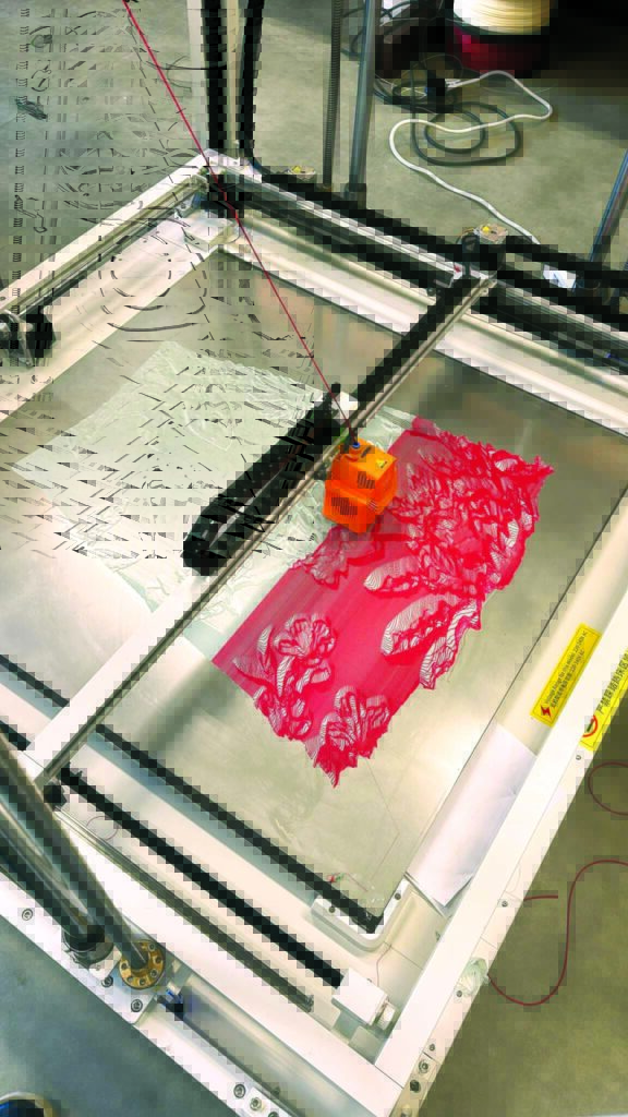 A 3D-printing arm on a fabrication platform creates intricate red lace fabric by Danit Peleg on a flat, metallic surface.