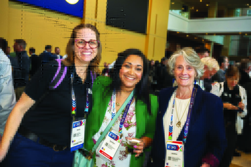 Three women, Robin DuBroy, formerly of Shade Sail University; Donielle Lorelli of National Industries for the Blind; and Andrea Lynn, of Sonoma Shade Sails, pose together at the Advanced Textiles Expo opening reception, smiling and wearing lanyards. The background features attendees and vibrant colors.
