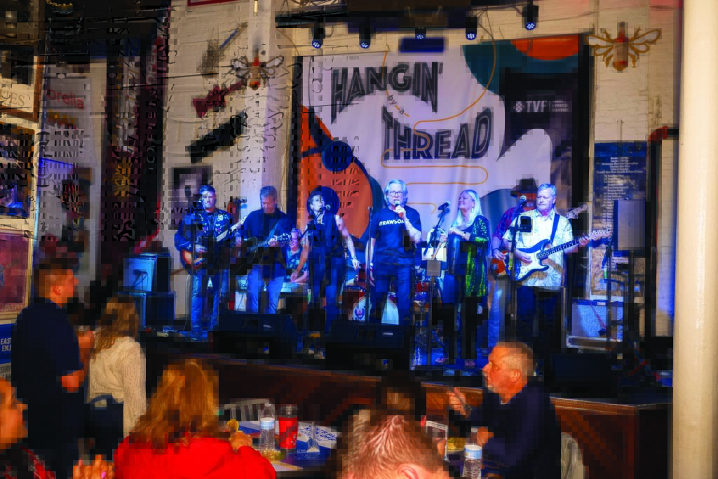 A lively performance Textile industry band Hangin' by a Thread performing on stage at Advanced Textiles Expo, featuring seven musicians, colorful backdrop and an engaged audience seated in foreground.