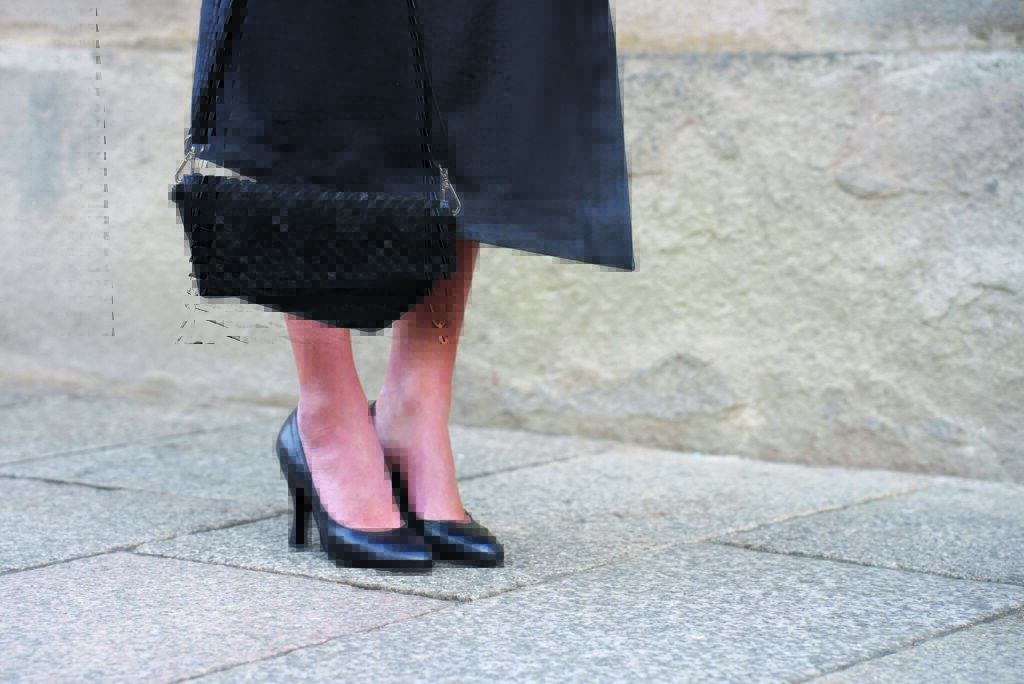 Stylish black high-heeled shoes paired with a textured black handbag, set against a light stone pavement backdrop.