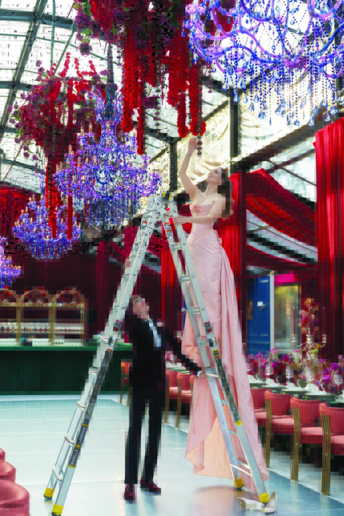 Bride Dakotah Terrace, owner of Jardiniere Events, in a pink gown and on a very tall ladder, adjusts a floral installation hanging from a chandelier. Groom Devin Van Bortel, owner of Amazing Event Rentals, wearing a tuxedo, holds the ladder in a lavish venue, a Horizon Series garden tent from Fred's Tents.