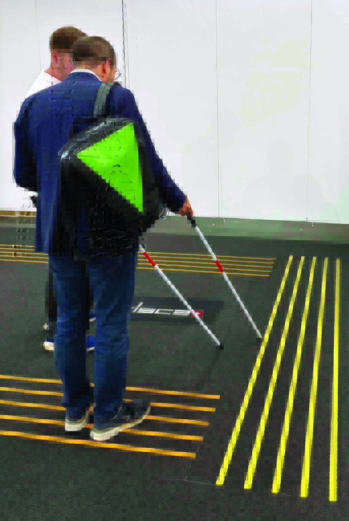 Two individuals stand on a textured floor with bright yellow tactile paving lines, one using a cane to navigate.