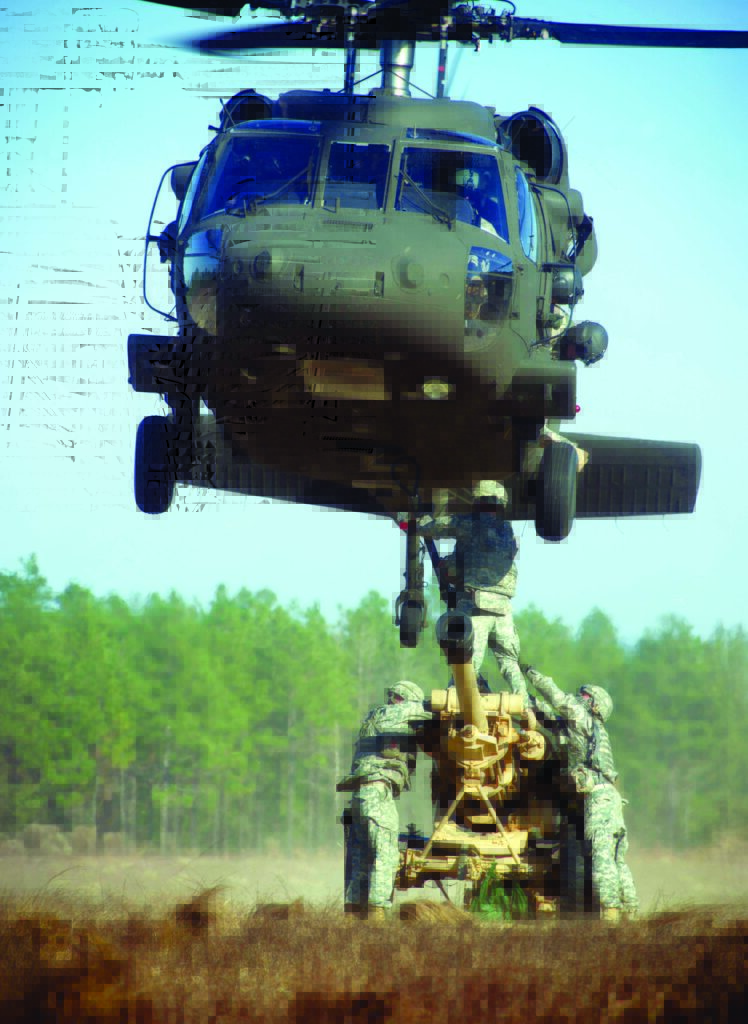 A UH-60 Blackhawk helicopter moves into position over a 105 mm howitzer. Soldiers are working in a green wooded area to sling-load the artillery piece.