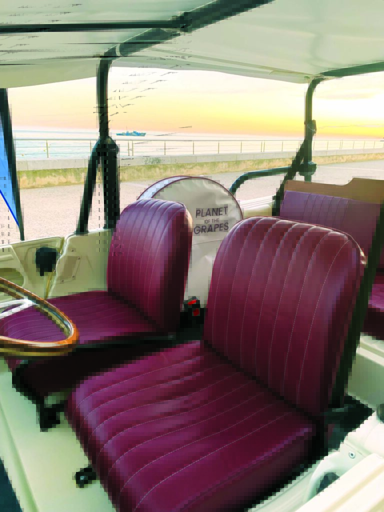 Two burgundy upholstered seats with white stitching inside a vehicle, featuring a circular sign that reads "PLANET OF THE GRAPES," overlooking a beach at sunset.