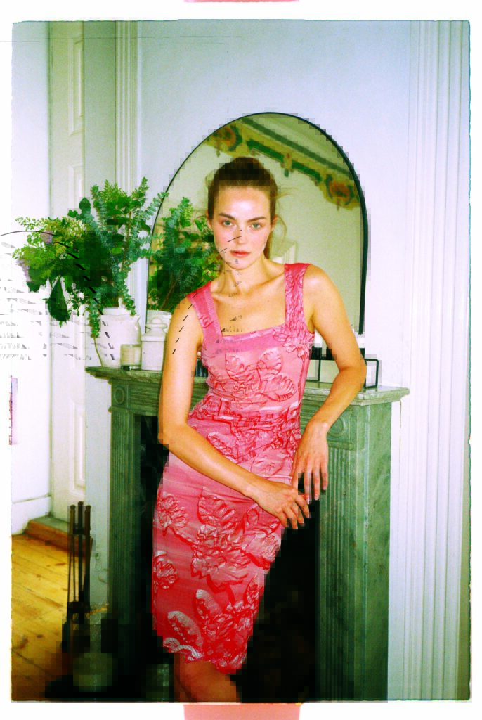 A woman in a pink floral dress made of 3D-printed fabric by Danit Peleg poses against a green marble fireplace, with plants and a mirror in the stylish interior background.