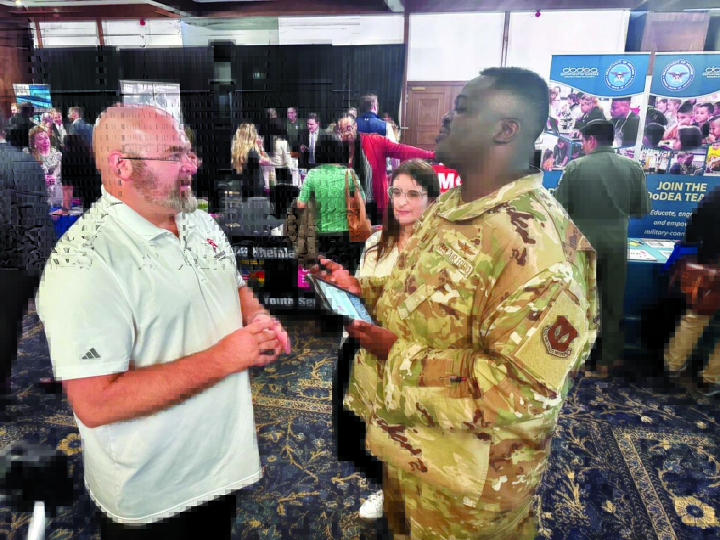 A military member in camouflage speaks with Steve Janke of Semper Forward, in a white polo shirt, at a workforce hiring event. Informational booths and attendees are in the background.