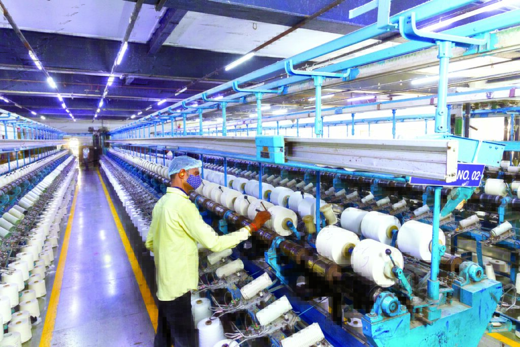 A textile worker operates machinery in a brightly lit factory, surrounded by rows of bobbins filled with white thread.