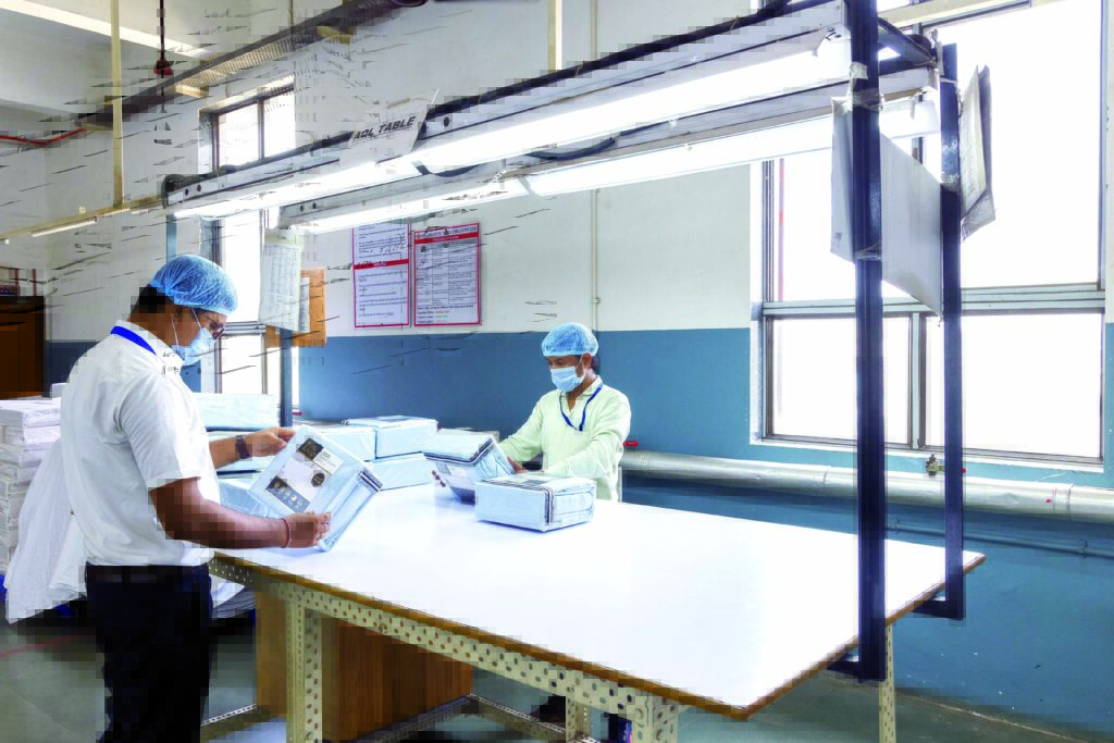 Two individuals in a textile factory setting, one reviewing documents and the other inspecting fabric packages, with bright overhead lighting.