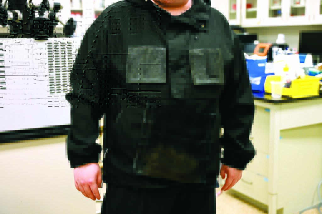 A person stands wearing a black jacket with two chest pockets and a transparent front pocket, surrounded by laboratory equipment.