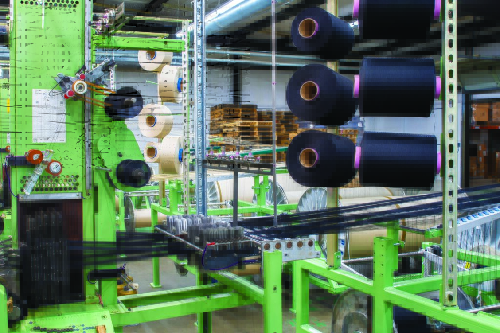 Green machinery is shown with black and cream spools of thread, surrounded by stacks of pallets in a brightly lit MMI Textiles factory.
