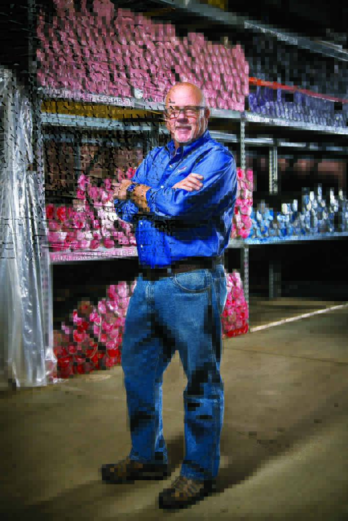 A man with glasses stands confidently with arms crossed in a warehouse, surrounded by colorful rows of natural fiber samples on shelves.