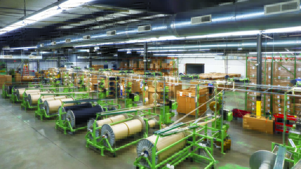 A spacious factory interior with green machinery holding large spools of fibers, with organized rows of cardboard boxes in the background. The 30,000-square-foot MMI Textiles Lenoir, N.C., narrow weaving plant opened in 2021.