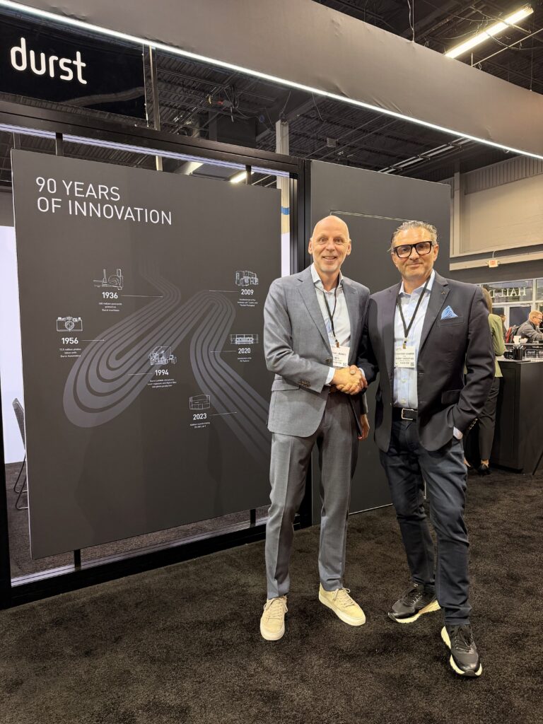 Two men shake hands in front of a black wall showcasing "90 Years of Innovation" with timeline graphics and white text.