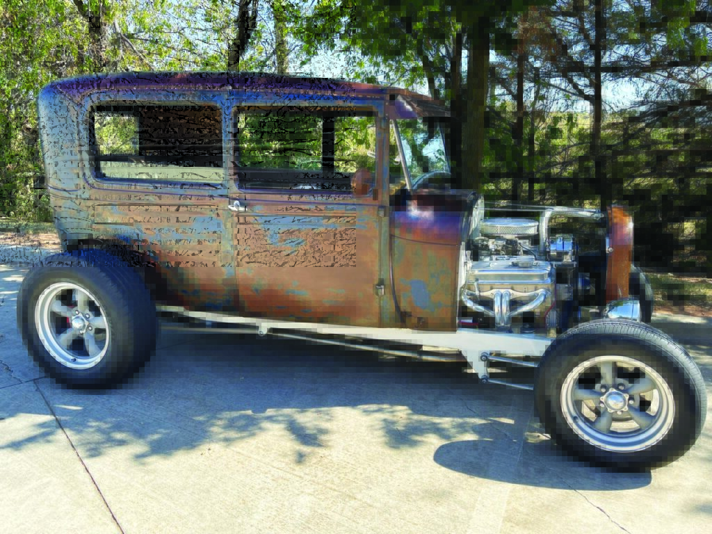Rustic Model A hot rod with a weathered patina, showcasing a modified engine and classic styling, parked under leafy trees.