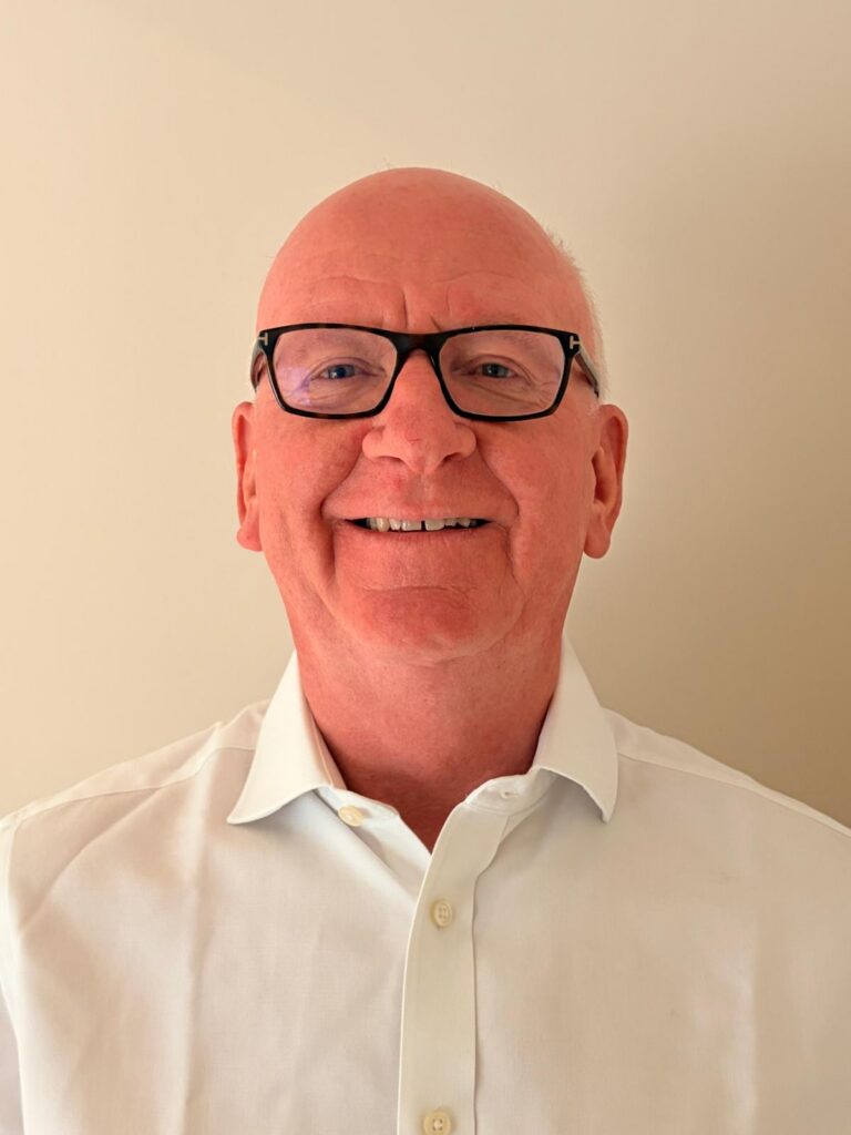 Smiling man with short, balding hair, wearing black glasses and a white collared shirt against a neutral beige background.