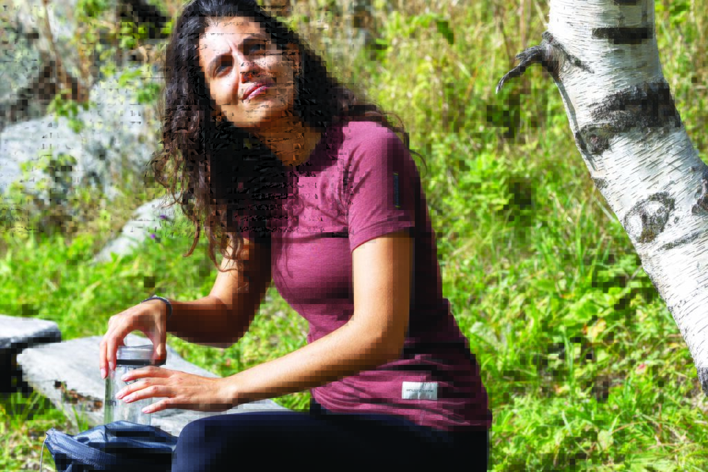 A woman in a maroon shirt sits outdoors next to a birch tree, holding a water bottle, with greenery and rocks in the background.
