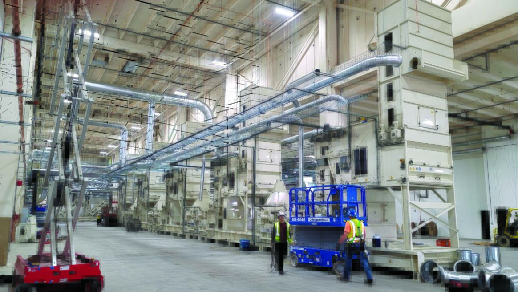 A spacious industrial facility with large machinery, ventilation ducts, and workers in safety gear moving around on a concrete floor.