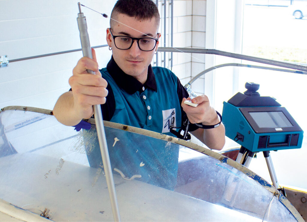 A person in a blue and black shirt holds a tool over a transparent surface, working with digital fabrication equipment nearby.