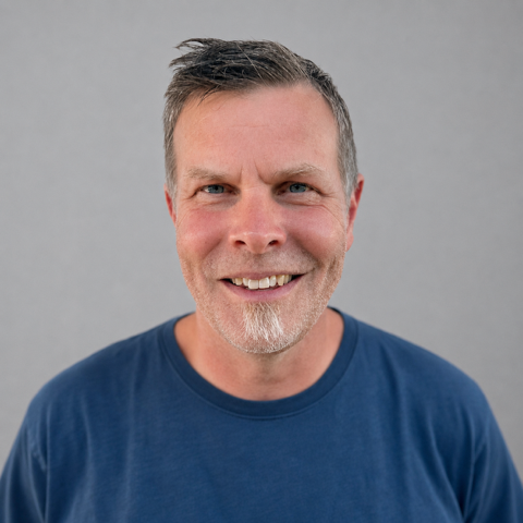 Schotex Smiling middle-aged man with short gray hair and a beard, wearing a blue t-shirt, against a light gray background.