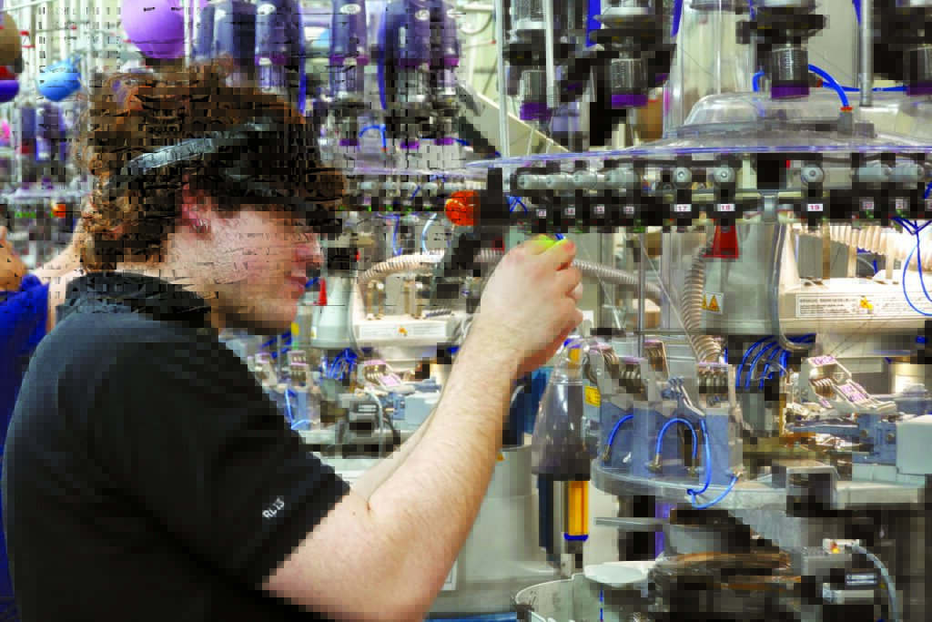 A knitting technician wearing a THREAD-X headset inspects machinery in a colorful textile factory, with various threads and automated equipment visible.