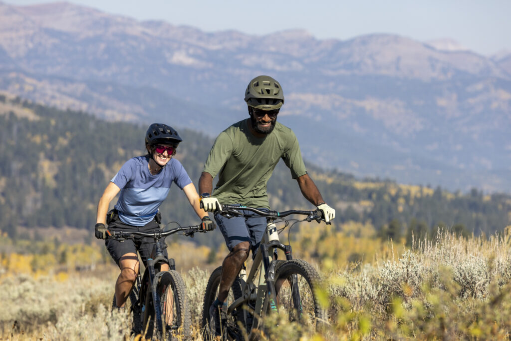 Two mountain bikers, a woman in a black helmet and a man in a green shirt, ride on a dusty trail with mountains in the background.