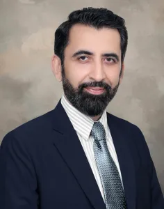 Portrait of a man with a beard, dressed in a dark suit and striped tie, smiling against a muted gray background.