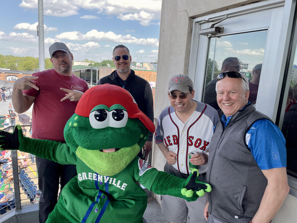 Five men pose with a green frog mascot wearing a red cap and a jersey. The background features a vibrant outdoor setting with a crowd.