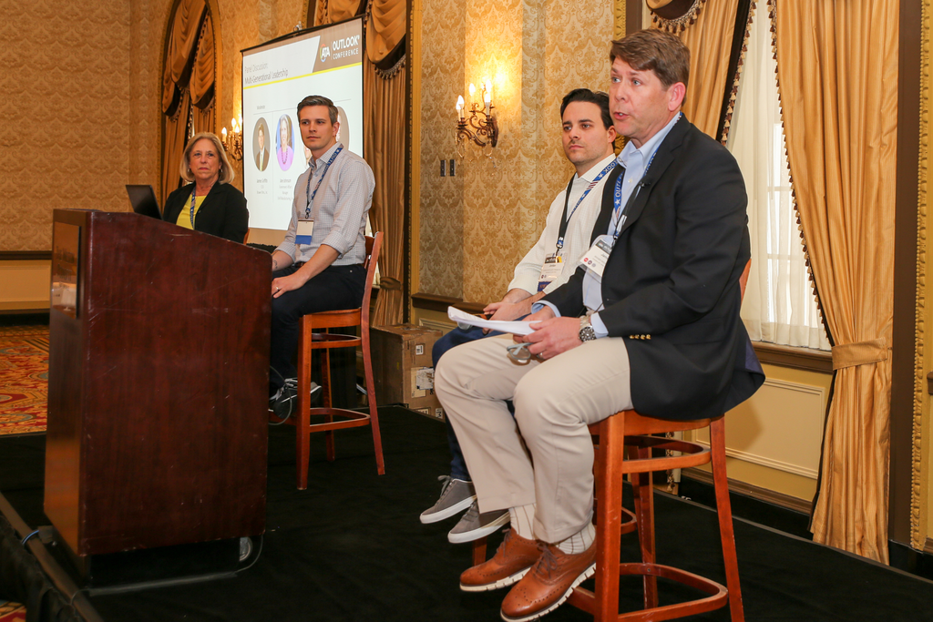 A panel discussion with four speakers; one at a podium and three seated, in a warmly lit room with beige walls and elegant curtains.