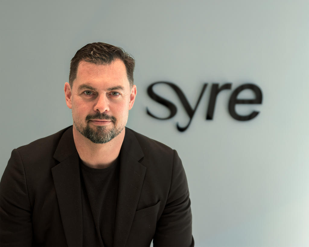 A man with a beard and short hair sits confidently, wearing a black jacket, in front of a grey wall with the word "syre" in a sleek font.