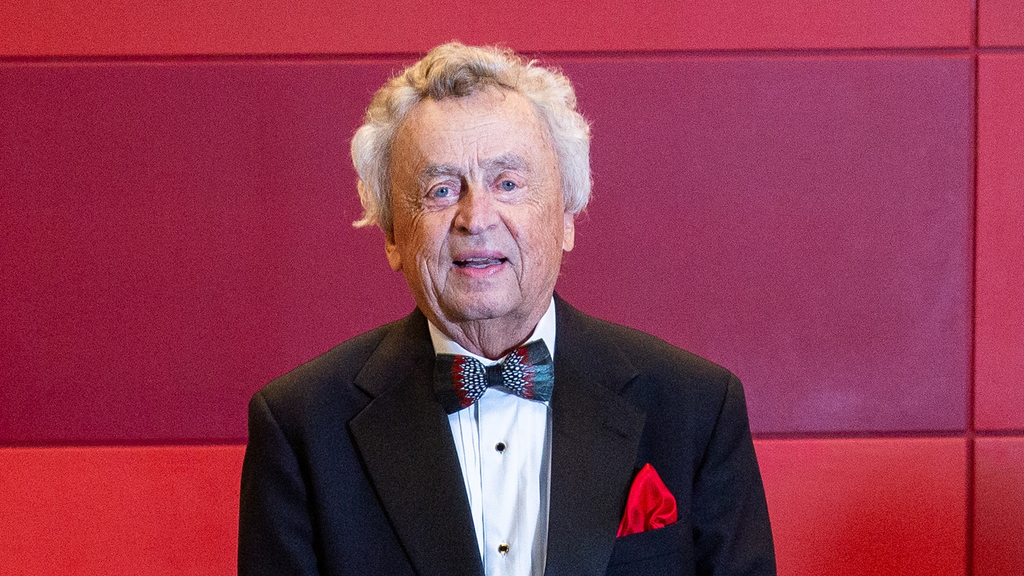Elderly man in a black tuxedo with a bow tie, smiling against a red and maroon background, wearing a red pocket square.