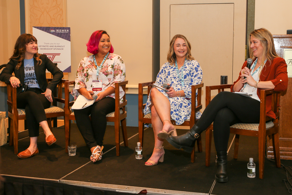 Four women seated on chairs at a panel discussion, smiling and engaged. The background features a podium and banners in soft colors.